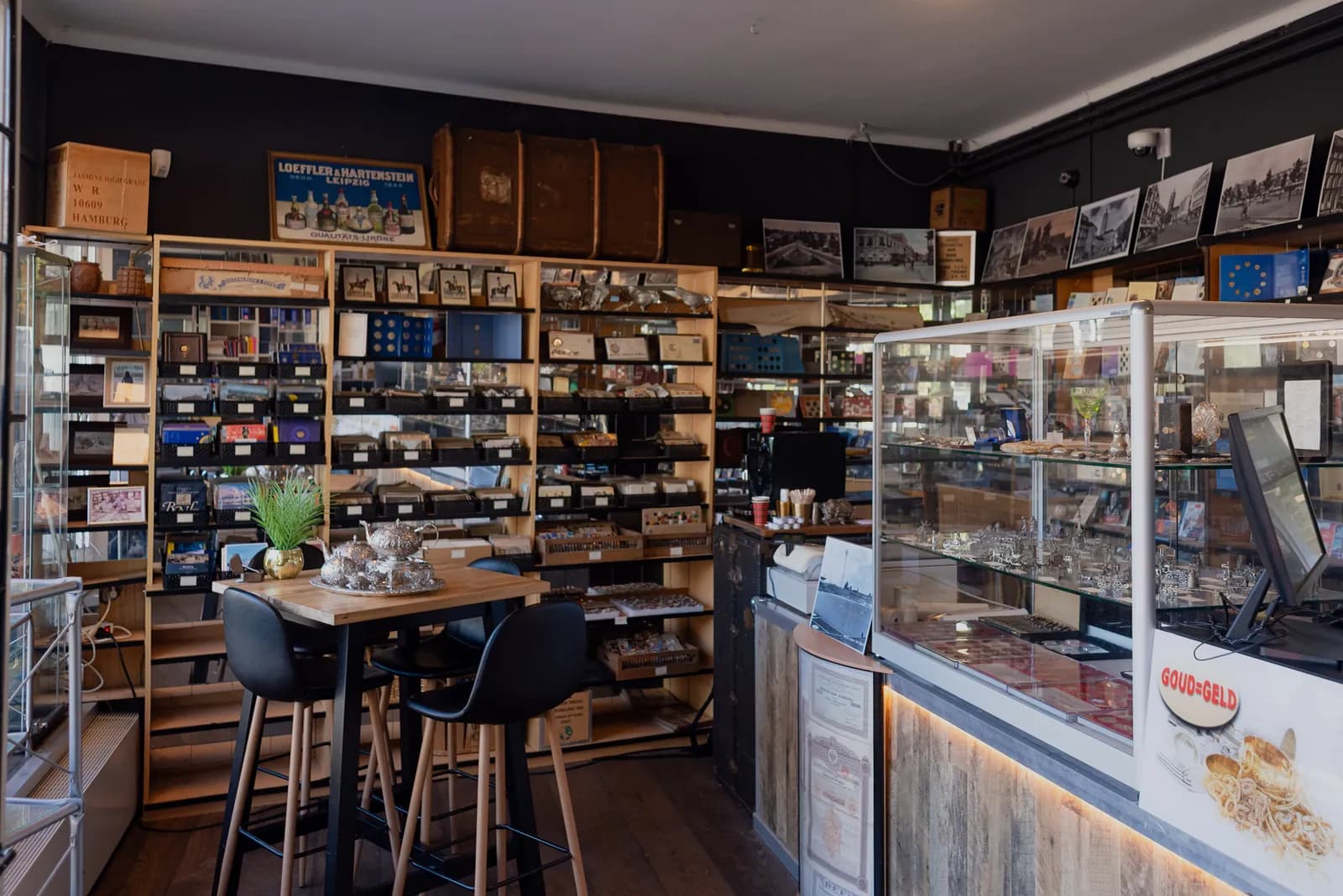 De koffiecorner in de winkel: een statafel met krukken en een zilveren theeservies, naast de toonbank met vitrine.